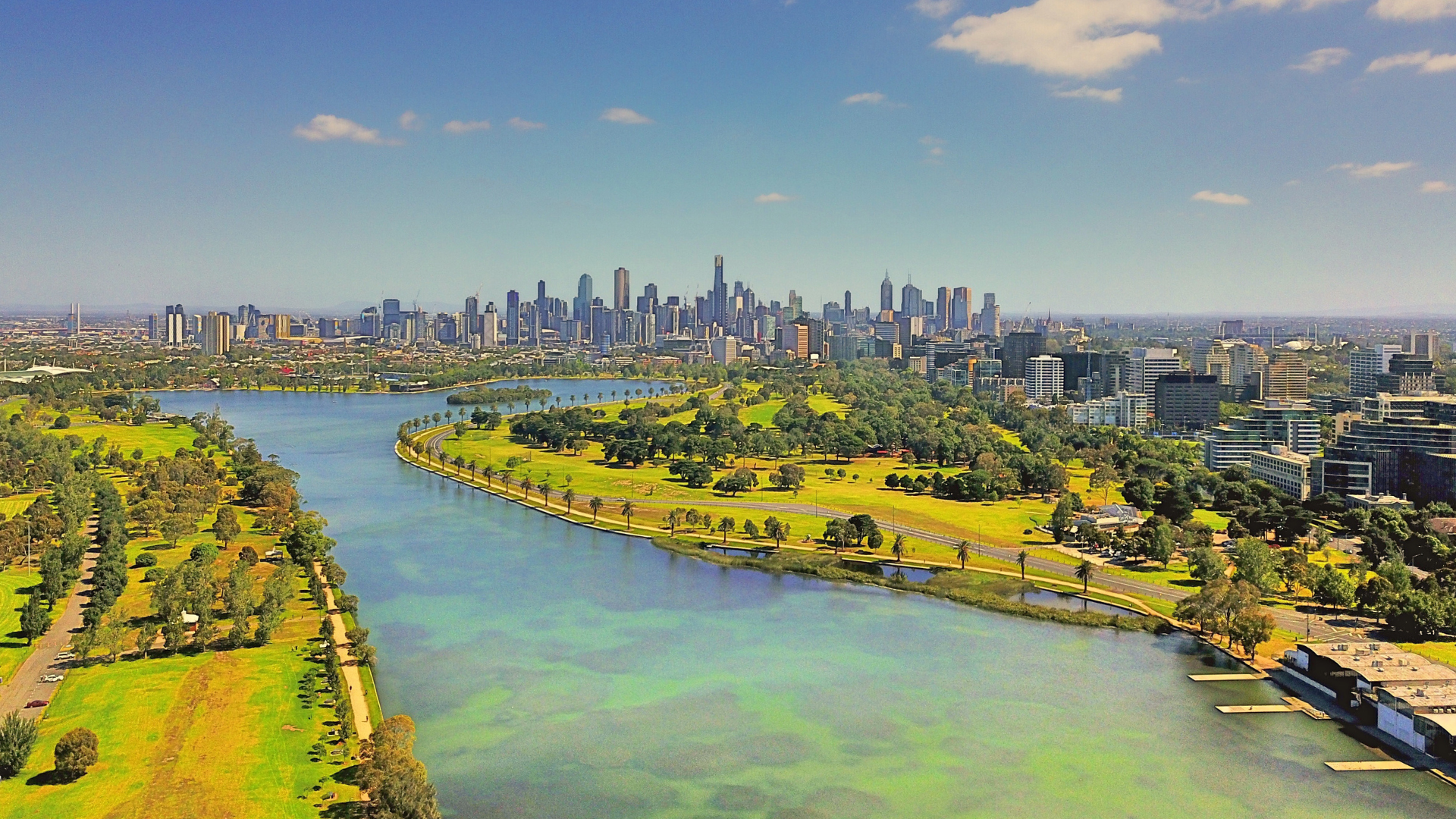 A tranquil lake from Albert Park Lane reflects the sunlight, with a modern cityscape of the Melbourne skyline rising majestically in the background.